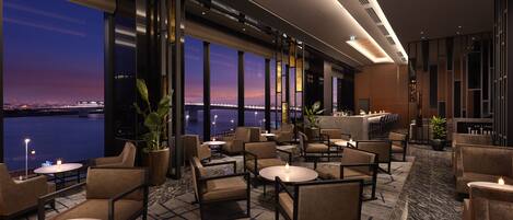 A modern hotel lobby with a view of the city skyline at dusk, featuring comfortable seating areas and a bar.