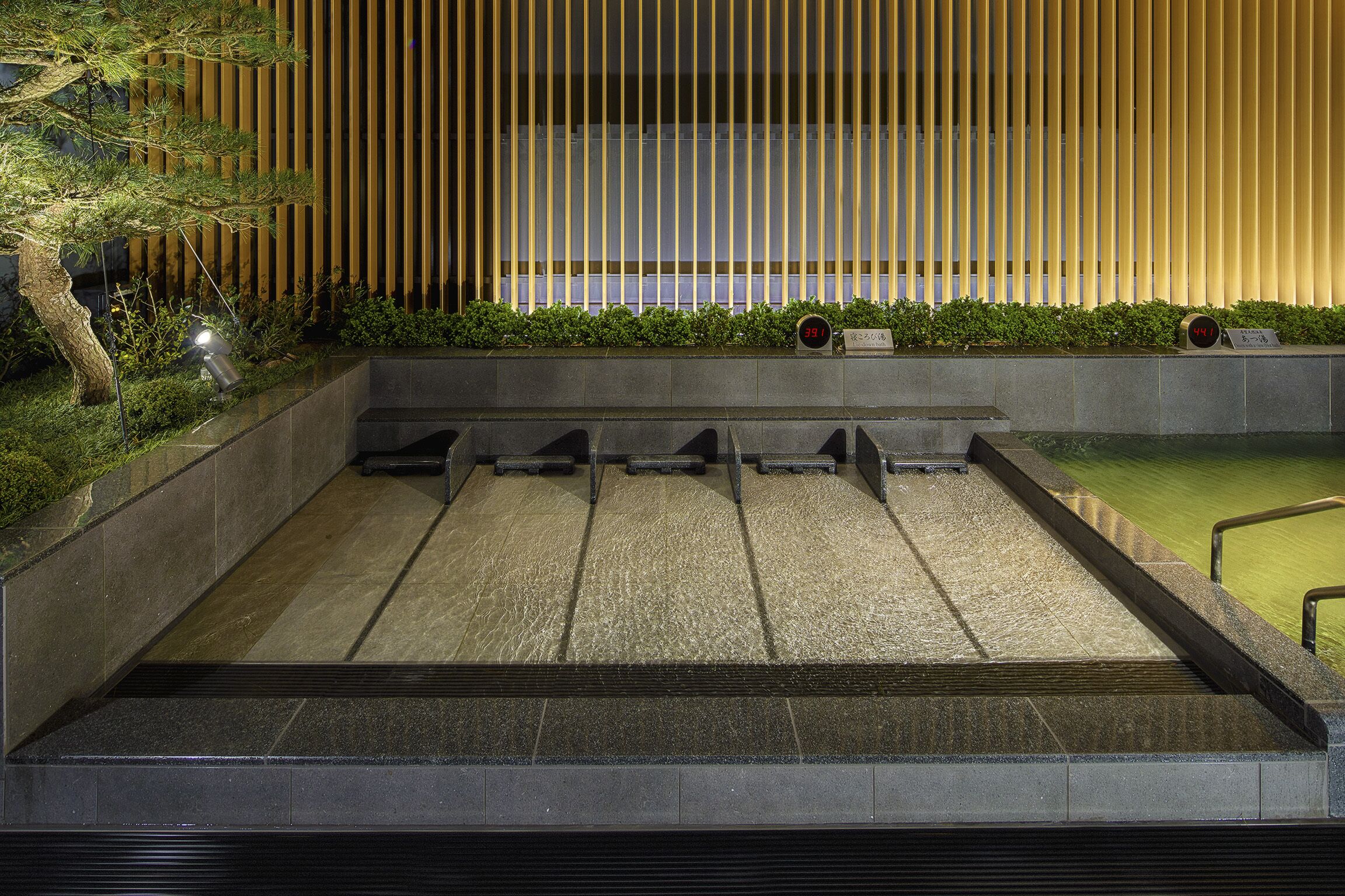 Un edificio moderno con una fuente de agua en primer plano, un muro de listones de madera y un reloj digital que muestra la hora.
