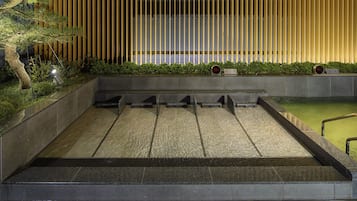 Un edificio moderno con una fuente de agua en primer plano, un muro de listones de madera y un reloj digital que muestra la hora.