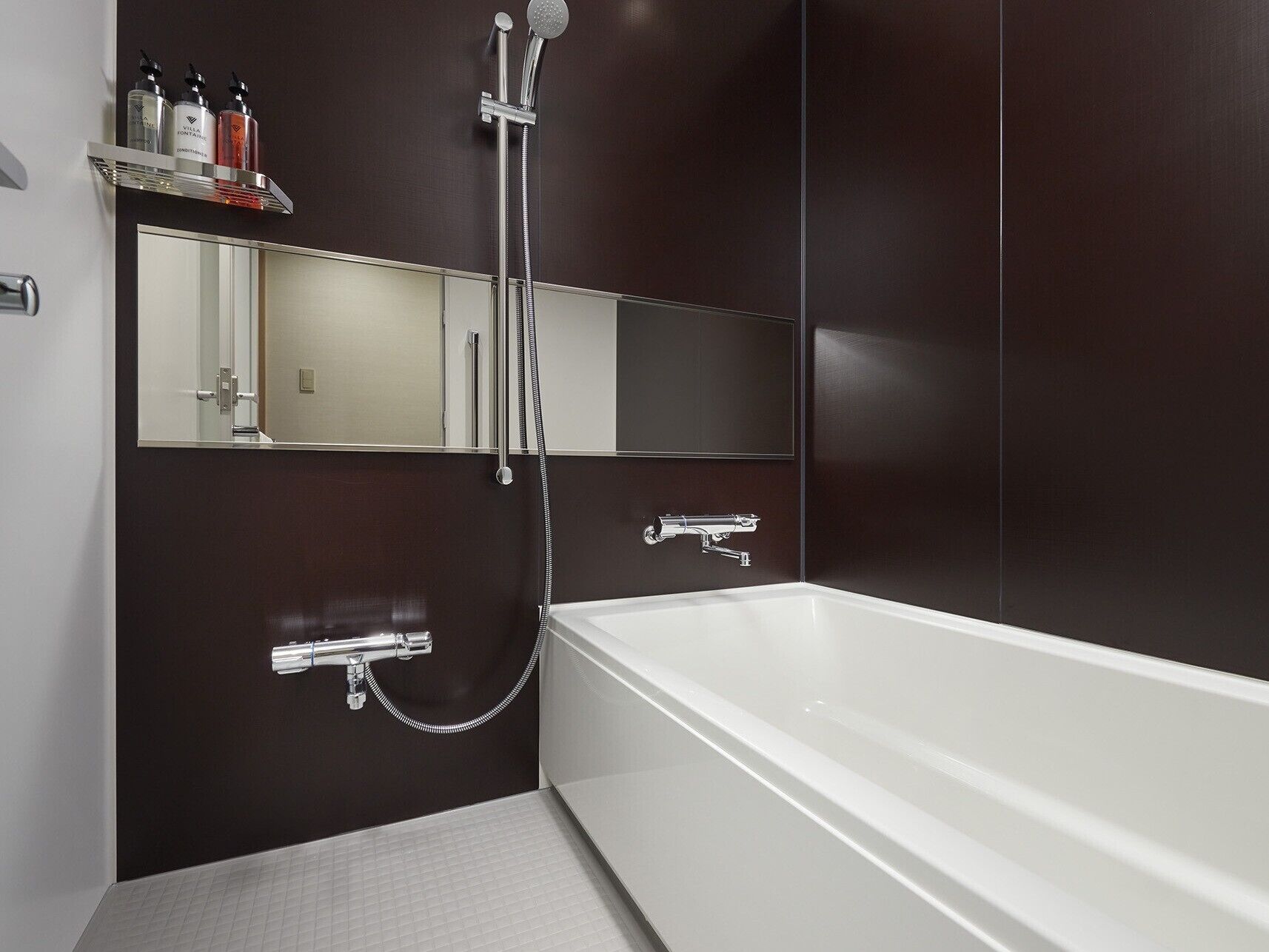 A modern bathroom with a large bathtub, a handheld showerhead, and a wall-mounted faucet. There is a shelf with toiletries and a mirror above the bathtub.