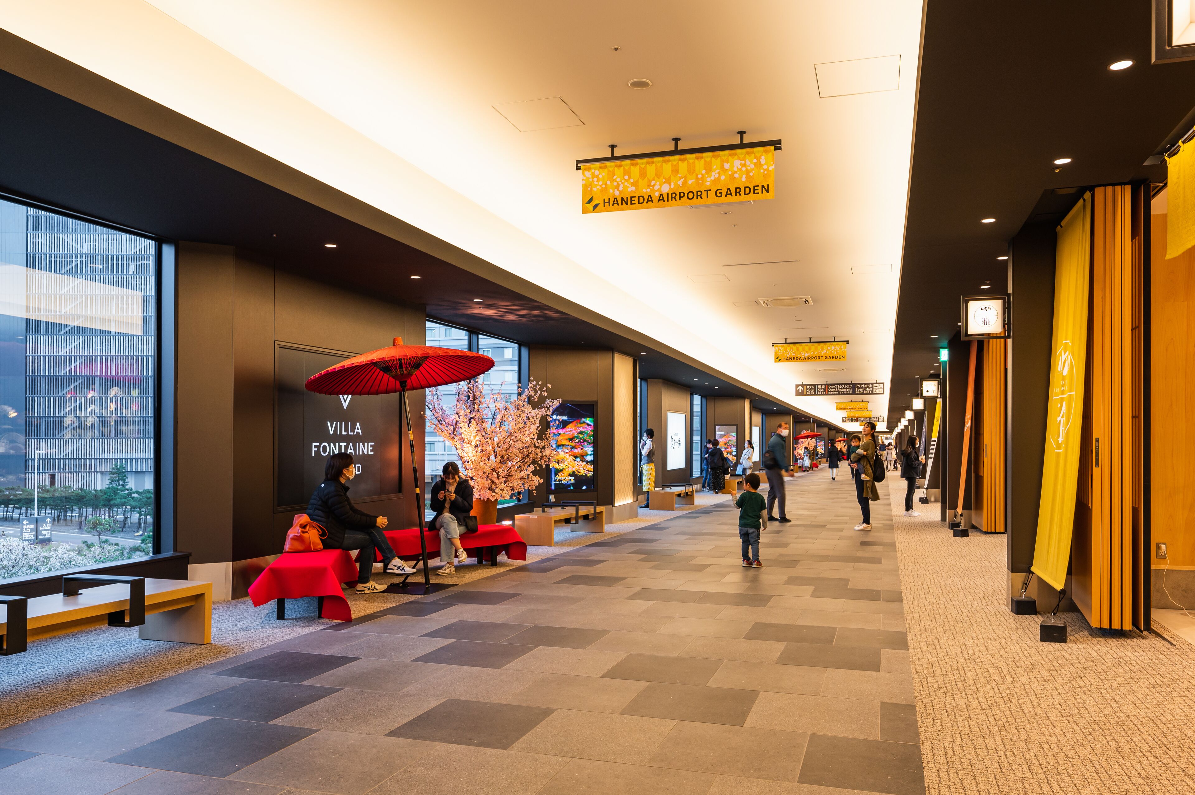 Ett modernt inomhusutrymme med bänkar och ett rött parasoll, en skylt med texten ”Villa Fontaine” och en banderoll med texten ”Haneda Airport Garden”.