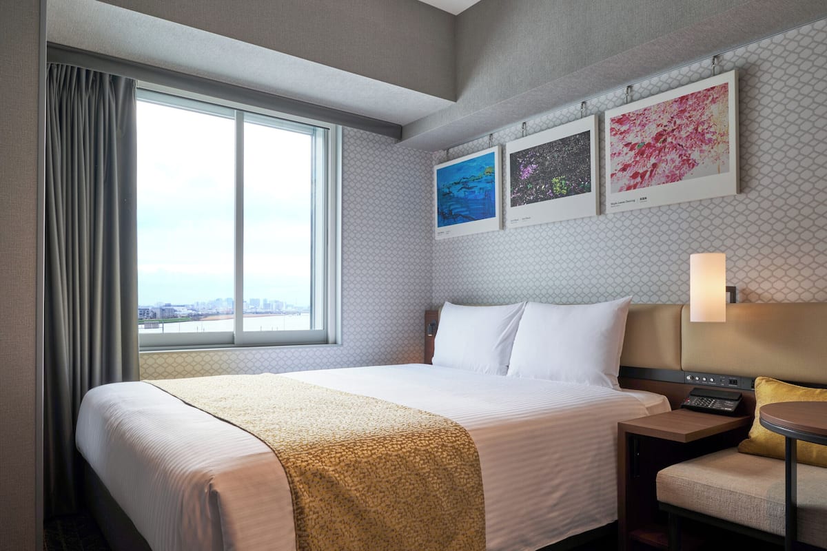 Ein Hotelzimmer mit einem großen Bett, einem Schreibtisch mit einem Laptop, einem Stuhl und einem Fenster mit Vorhängen. An der Wand hängen drei gerahmte Bilder.