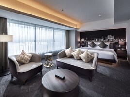Habitación de hotel moderna con una cama grande, dos sillones, una mesita redonda y vistas a la ciudad.