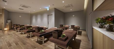 A spa room with multiple massage tables and chairs, wooden flooring, and a reception desk with a vase of flowers.