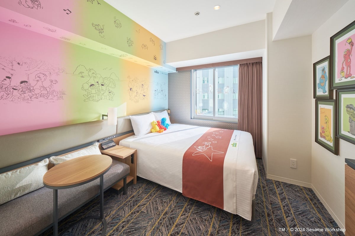 Ein Hotelzimmer mit Bett, Schreibtisch, Stuhl, einem Fenster mit Vorhängen und einer Wand mit Zeichnungen.