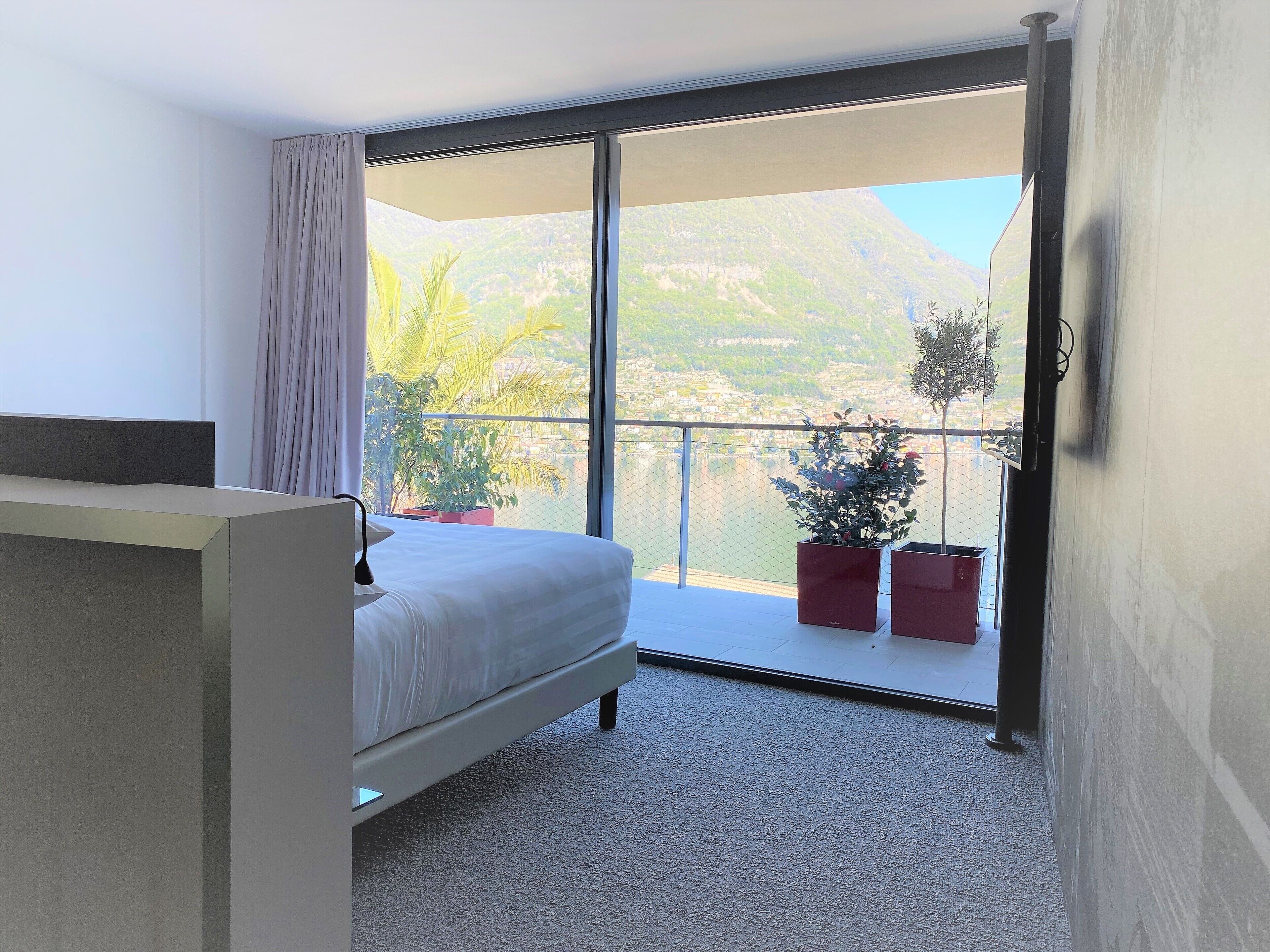 A bedroom with a bed, a large window, and a balcony with potted plants.