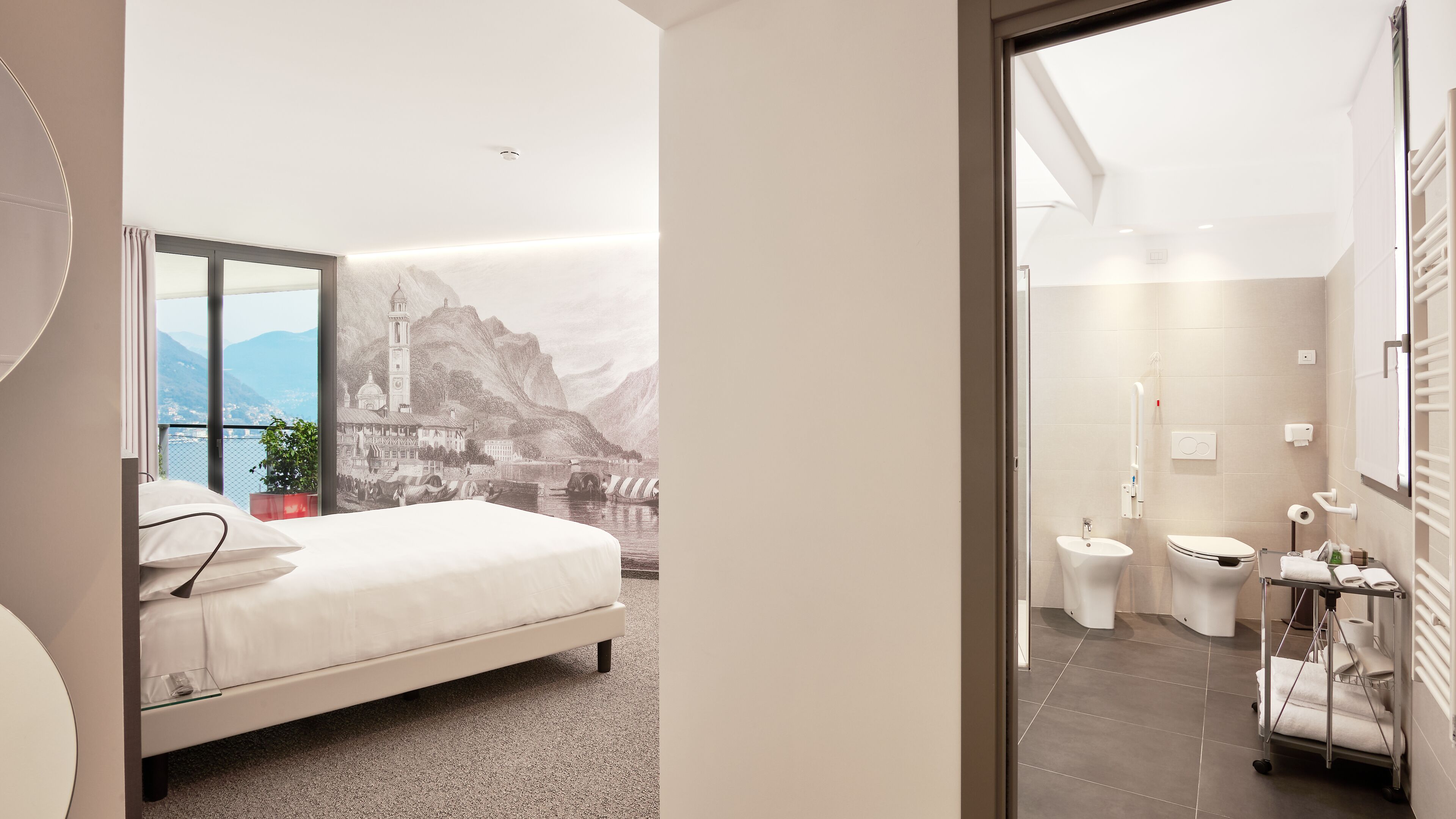 A bedroom with a bed, a view of a mountainous landscape, and a bathroom with a toilet, sink, and towel rack.