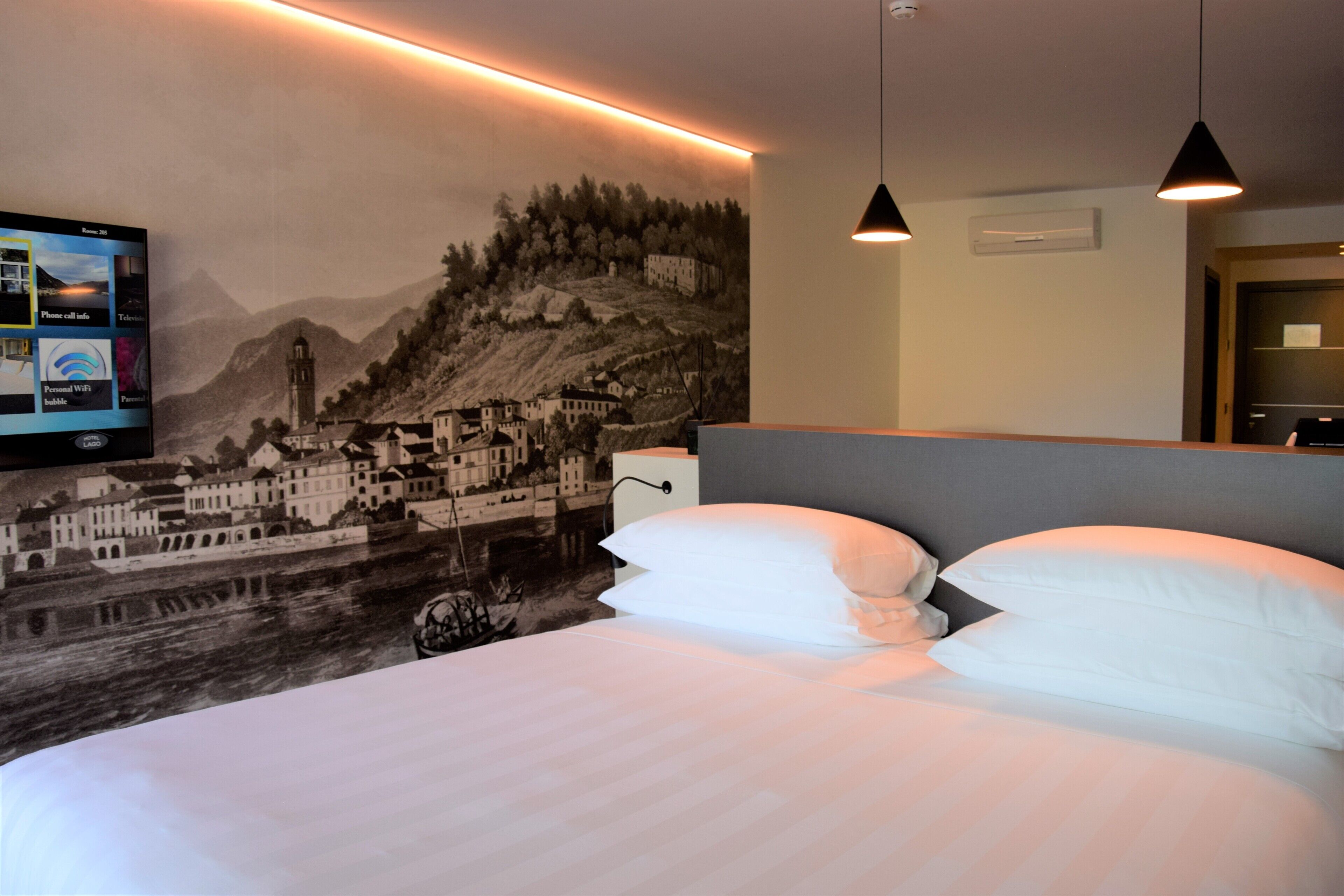 Quarto de hotel com cama, almofadas brancas, televisão fixada exibindo informações do quarto e um mural de uma cidade na parede.
