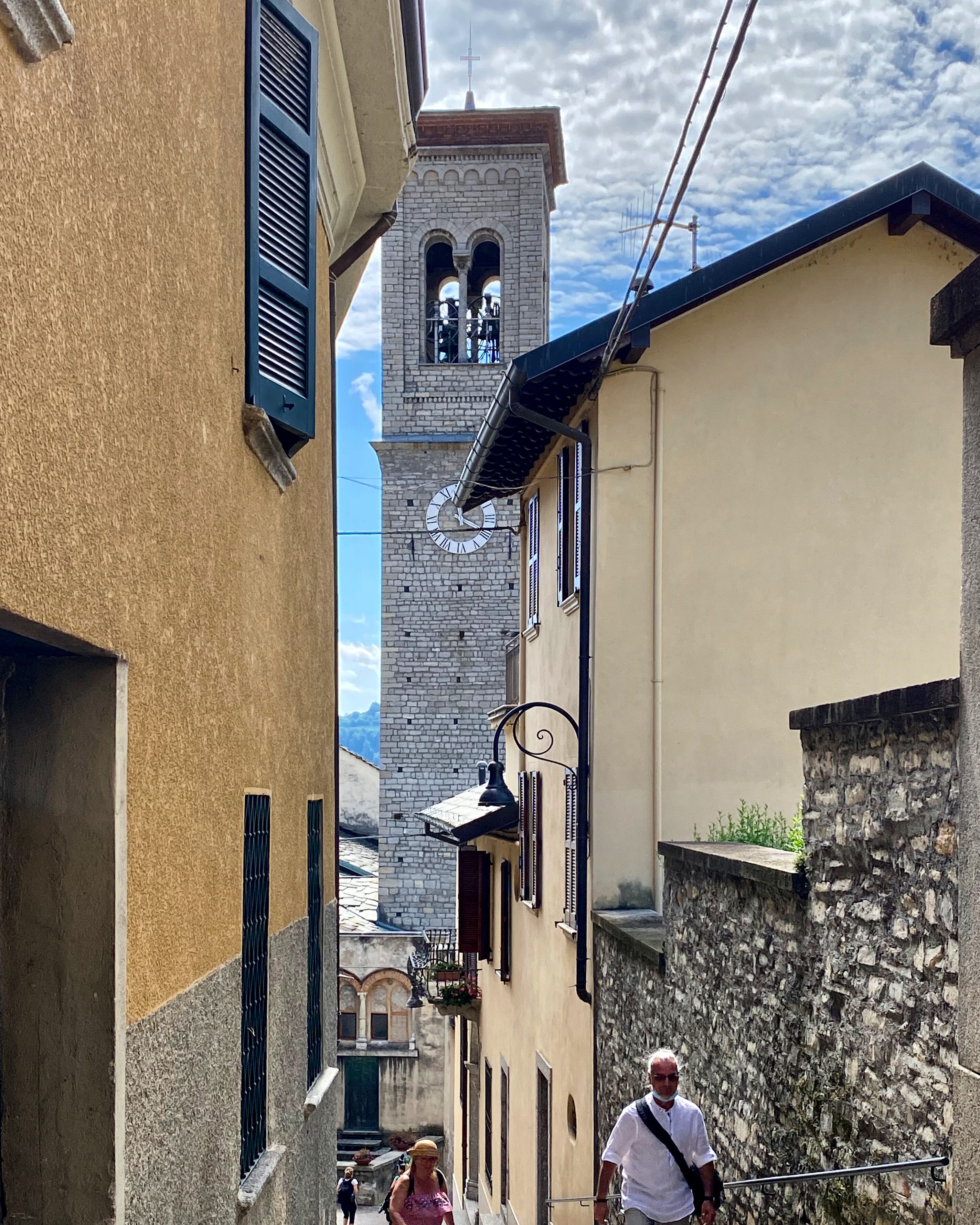 A narrow street with buildings on either side, a clock tower, and a cross on top.