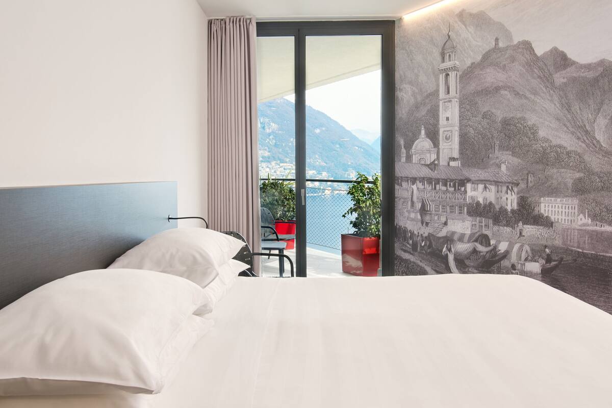Habitación de hotel con una cama grande, vista a una montaña y una masa de agua a través de un vidrio corredero, y un mural de un edificio histórico.