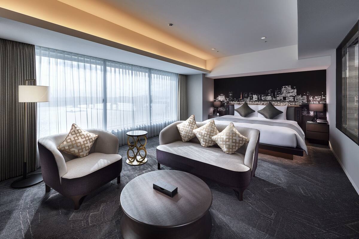 A modern hotel room with a large bed, two sofas, a coffee table, and a cityscape view.