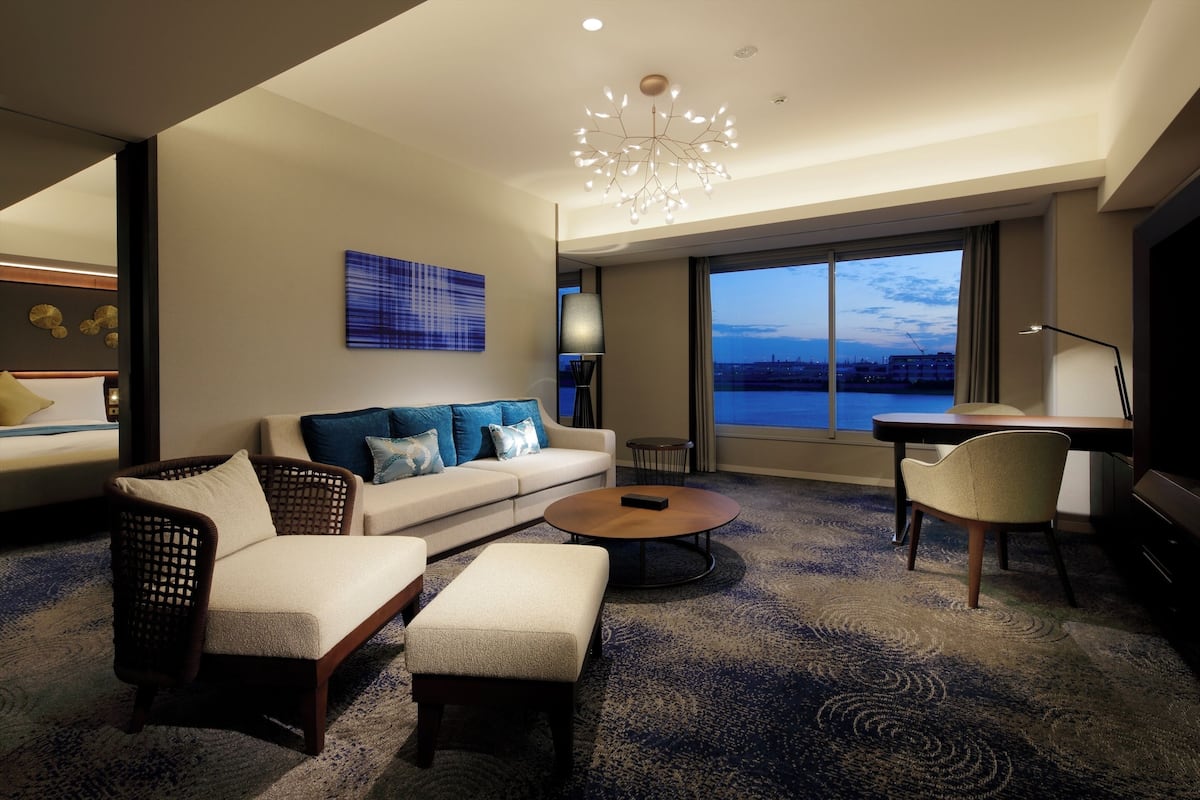 A modern hotel room with a large window, a sofa with blue cushions, a coffee table, and a dining area with a lamp.