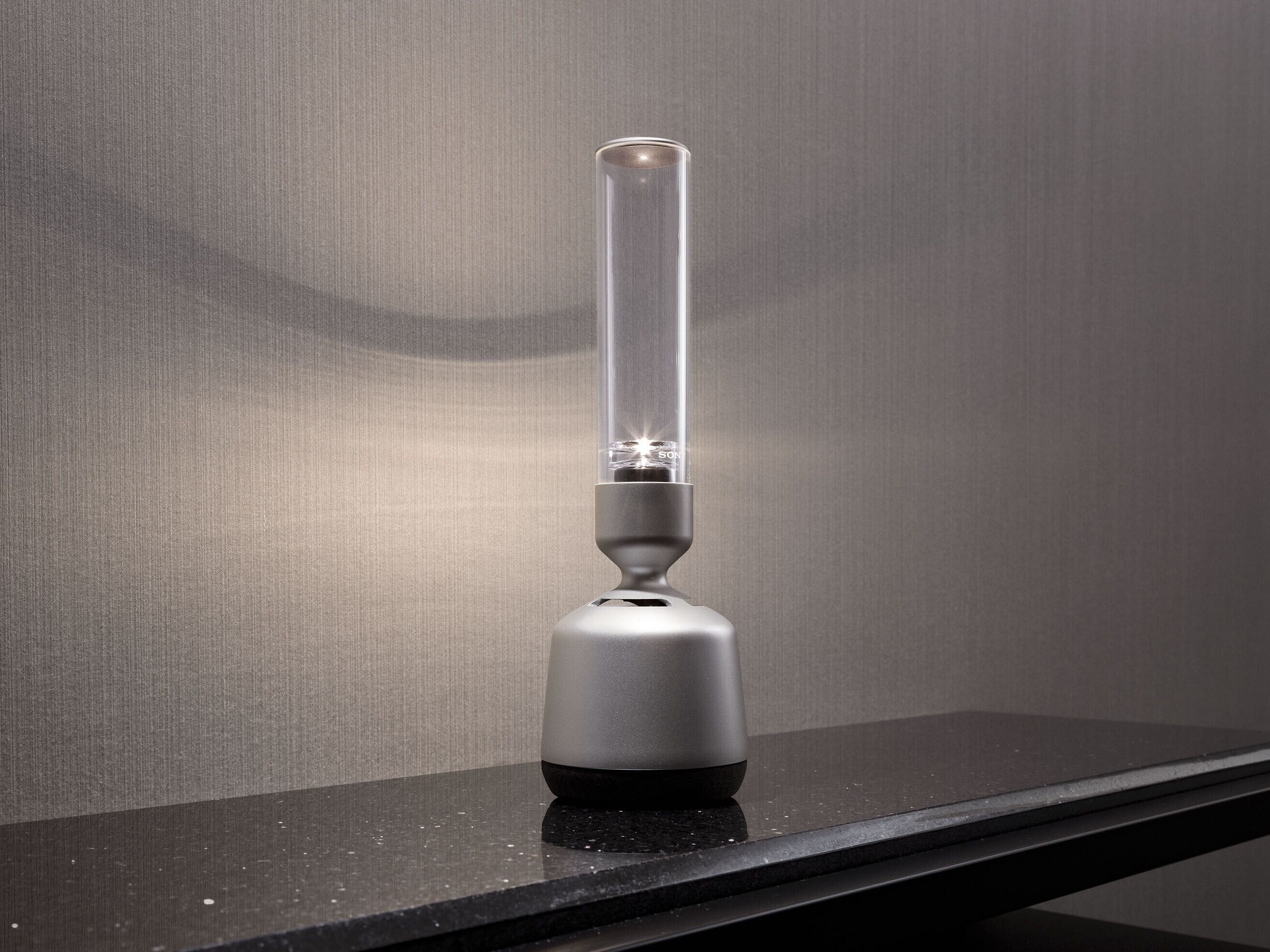 A modern, minimalist lamp with a clear glass enclosure and a metallic base on a dark countertop.
