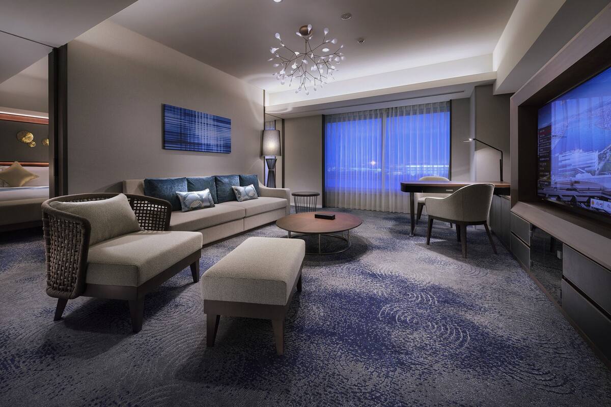 A modern hotel room with a sofa, ottoman, coffee table, and a TV screen displaying a cityscape.