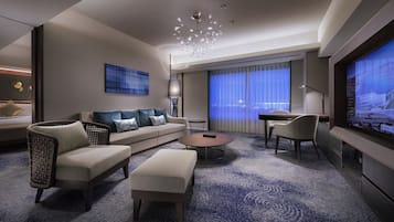 A modern hotel room with a sofa, ottoman, coffee table, and a TV screen displaying a cityscape.