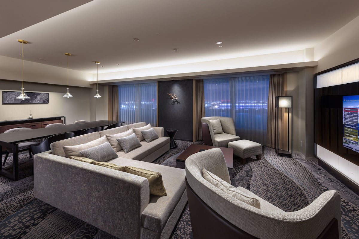 A modern hotel room with a large sofa, a smaller sofa, a chair, a desk, and a TV screen.