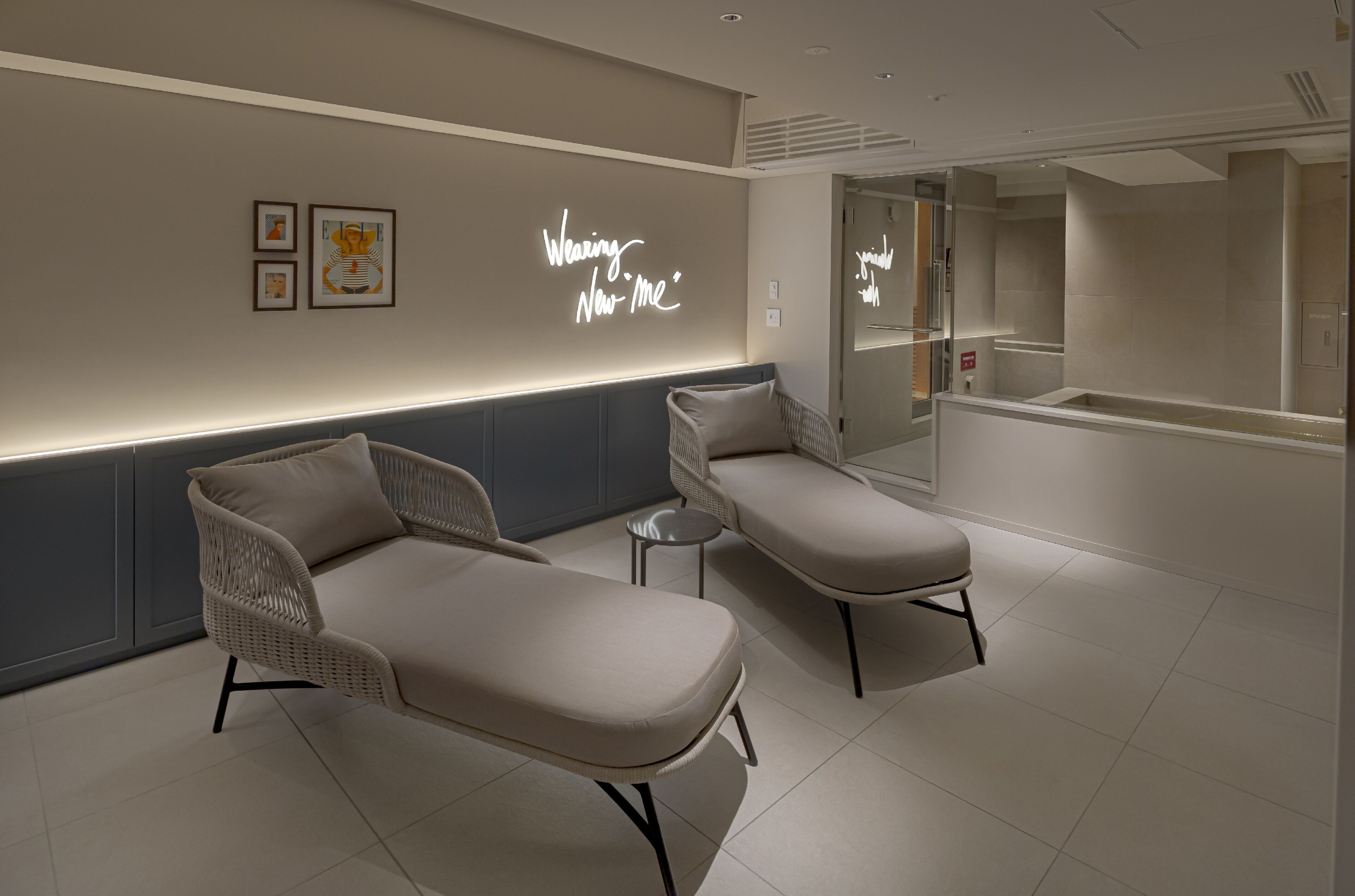 A modern spa room with two lounge chairs, a small round table, and a wall with framed artwork and a neon sign.
