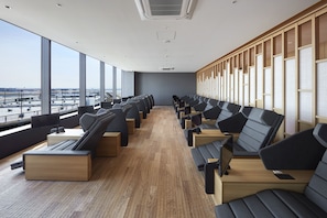 A modern hotel room with a long row of black leather sofas and wooden tables, large windows offering a view of the airport, and a wooden partition wall.