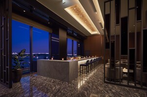 Zona bar moderna con pavimento in marmo, ampie finestre e vista sulla città al crepuscolo.