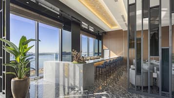 Een moderne bar met uitzicht op het water, een grote potplant en een zithoek met tafels en stoelen.