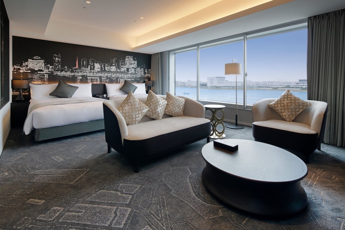 A modern hotel room with a large bed, two sofas, a coffee table, and a cityscape view through large windows.