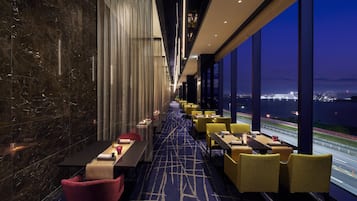 A hotel corridor with tables and chairs, large windows with a view of a cityscape and a body of water, and a patterned carpet.