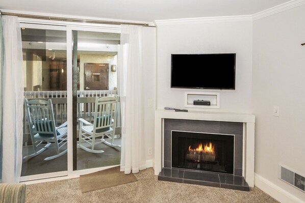 A living room with a fireplace, a television mounted above it, and a sliding glass door leading to a balcony with outdoor furniture.