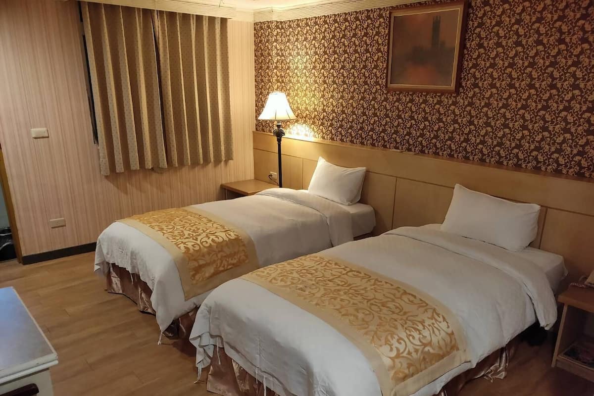 Habitació d'hotel amb dos llits, una tauleta de nit amb una làmpada, un terra de fusta i una paret amb un disseny.