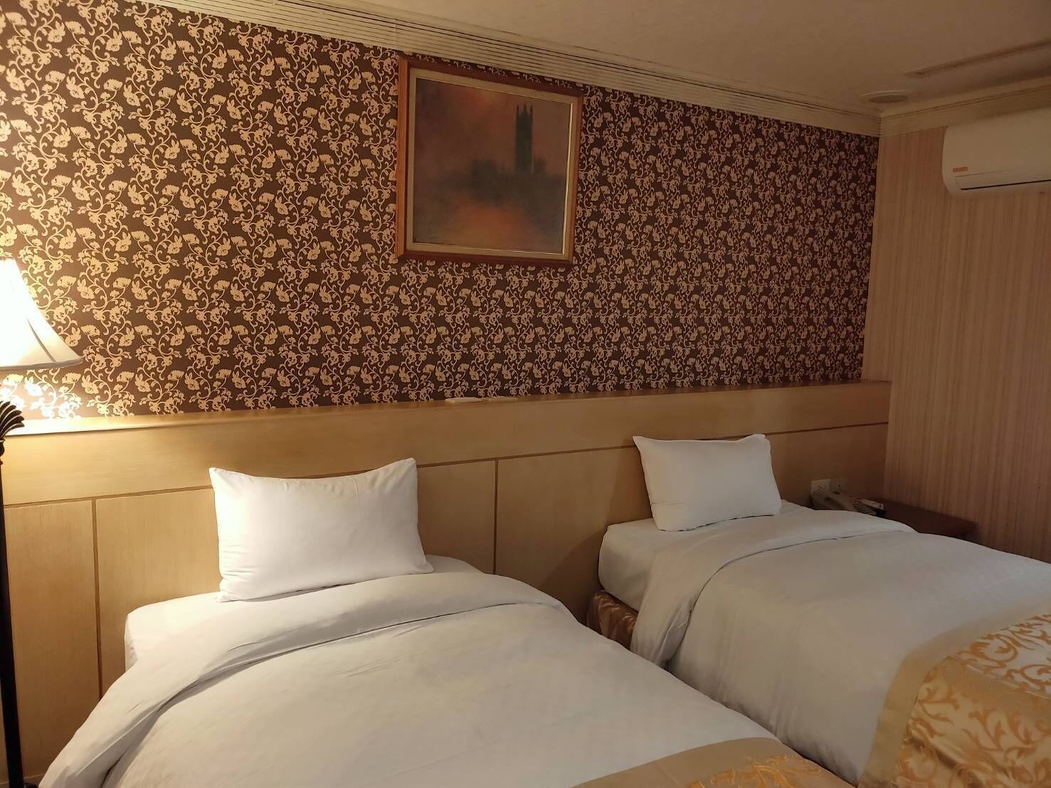 A hotel room with two beds, a painting on the wall, and an air conditioning unit.