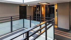 Een moderne hotel lobby met een glazen vloer die leidt naar een lager gelegen niveau, een receptiebalie en een lift.