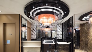 Een moderne hotellobby met een centraal gelegen receptiebalie, een grote kroonluchter en behang met patronen.