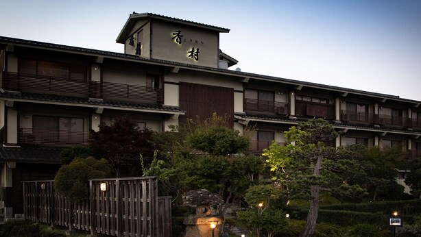 Un edificio tradicional japonés con varios pisos, barandillas de madera y un jardín bien cuidado con árboles y una formación rocosa.