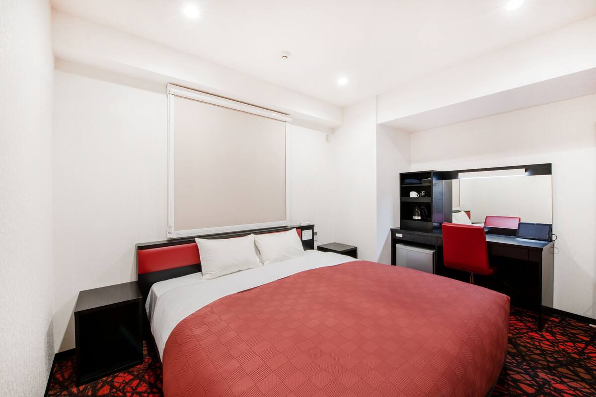 A hotel room with a large bed, a desk with a chair, a mirror, and a small kitchenette.
