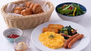 Prato de pequeno-almoço com ovos mexidos, salsichas e vegetais, uma salada numa tigela, uma cesta de croissants e um pequeno recipiente de compota.