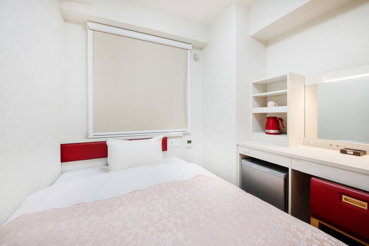 A hotel room with a bed, a red headboard, a white bedside table, a red kettle, a white coffee maker, a white sink, and a mirror.