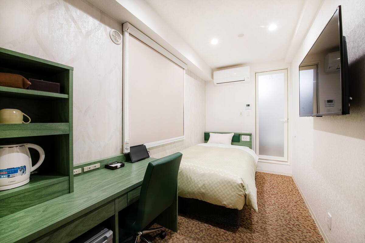 A hotel room with a bed, a desk with a chair, a television, and a window with blinds.
