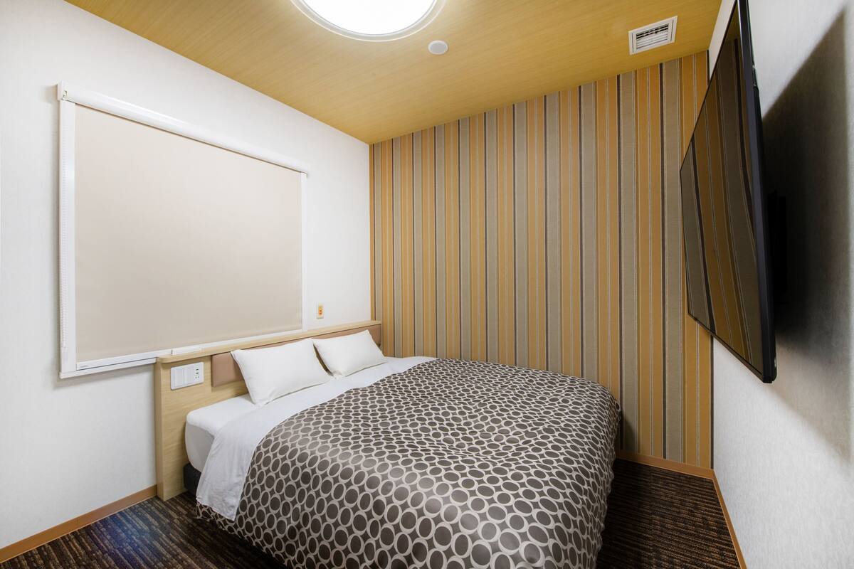 A hotel room with a bed, a large window, a wall-mounted TV, and a striped wall.