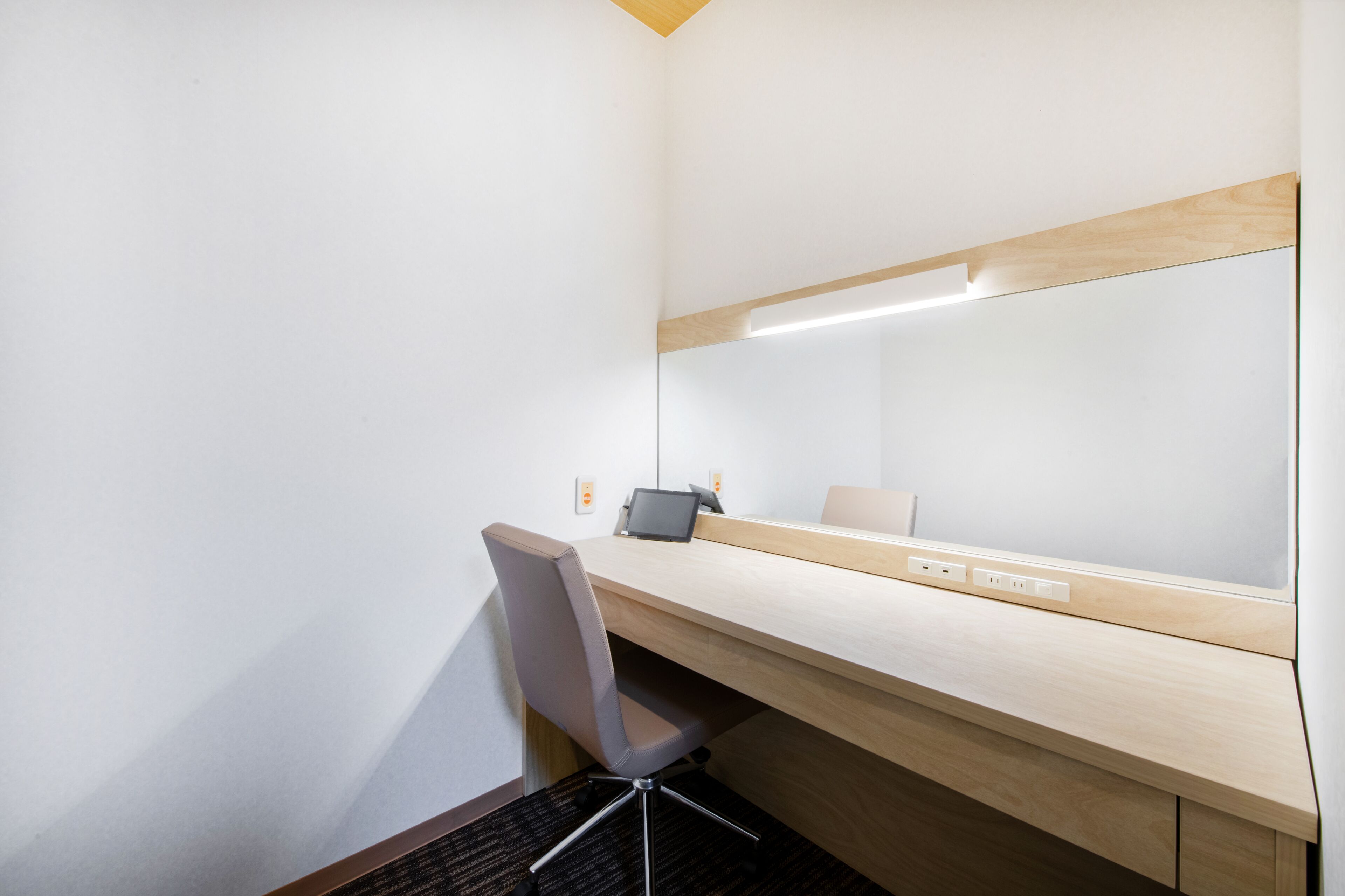A modern hotel room with a wooden desk, a chair, a large mirror, and a tablet.