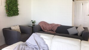 A living room with a white couch, a grey couch, and a white armchair. There is a small table with a plant and a pink blanket.