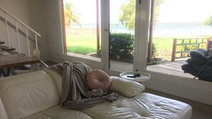 A white leather sofa with a blanket and pillows, a small white table with a remote control, and a view of a beach and palm trees through large windows.