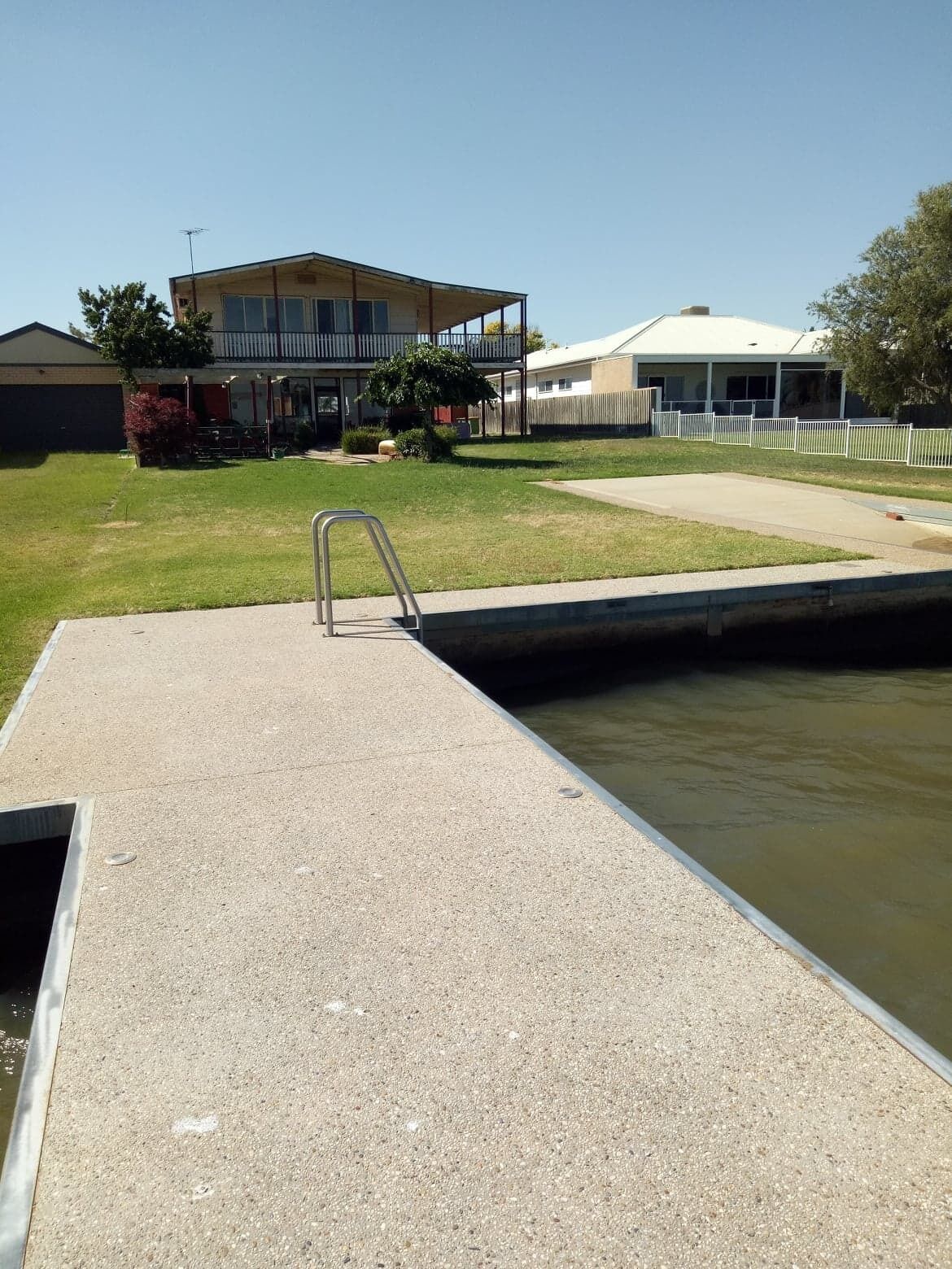 A swimming pool with a metal ladder, surrounded by a concrete deck. In the background, there is a two-story building with a balcony and a covered area.