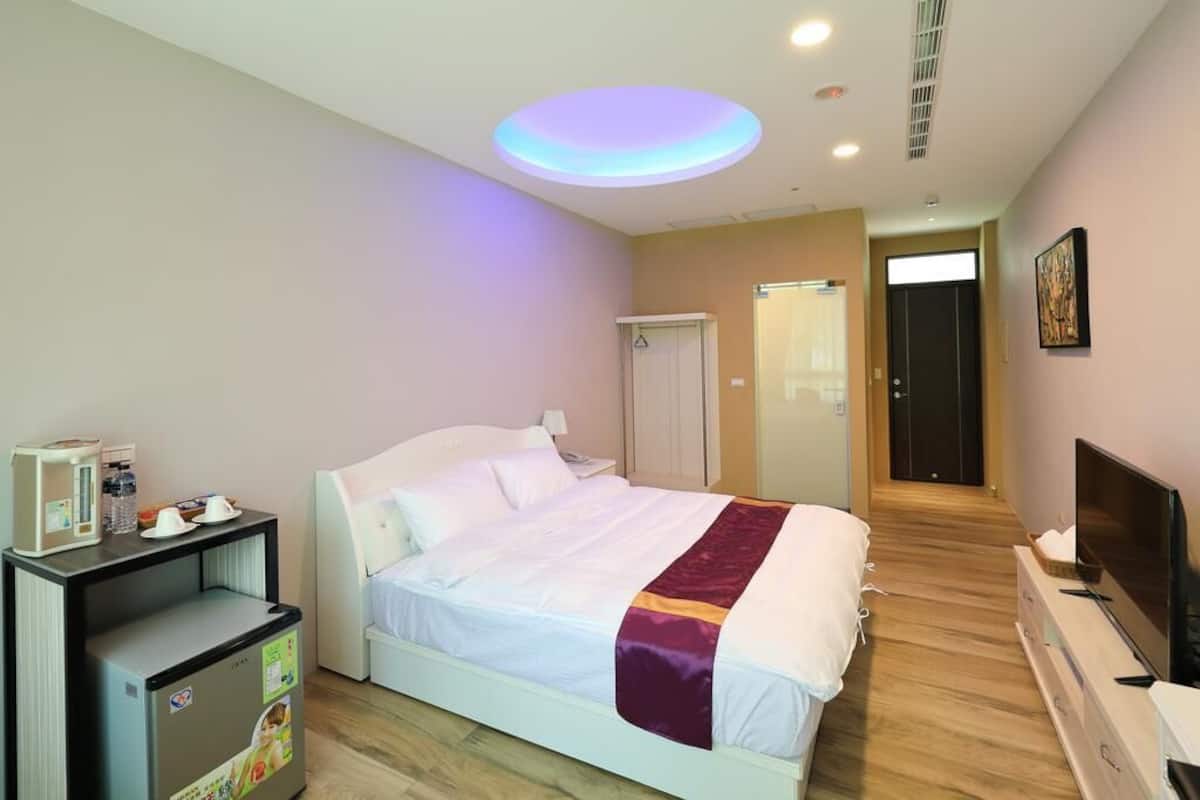 A hotel room with a bed, a desk with a refrigerator, a television, and a door leading to the bathroom.