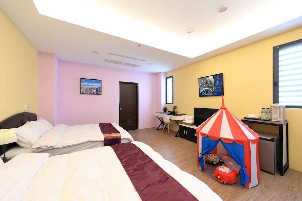 A hotel room with two beds, a desk, a chair, a small tent, and a refrigerator.