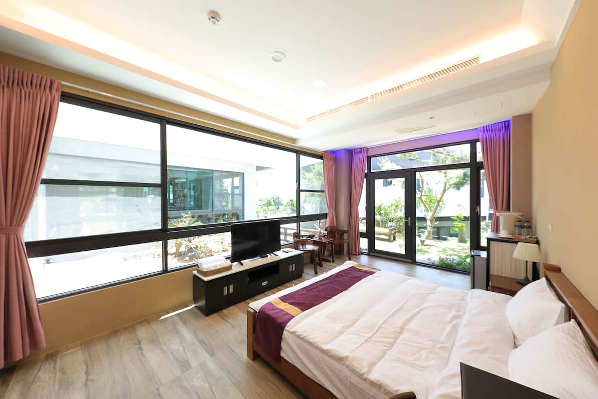 A modern hotel room with a large bed, a flat-screen TV, a desk, and a view of the outdoors through large windows.