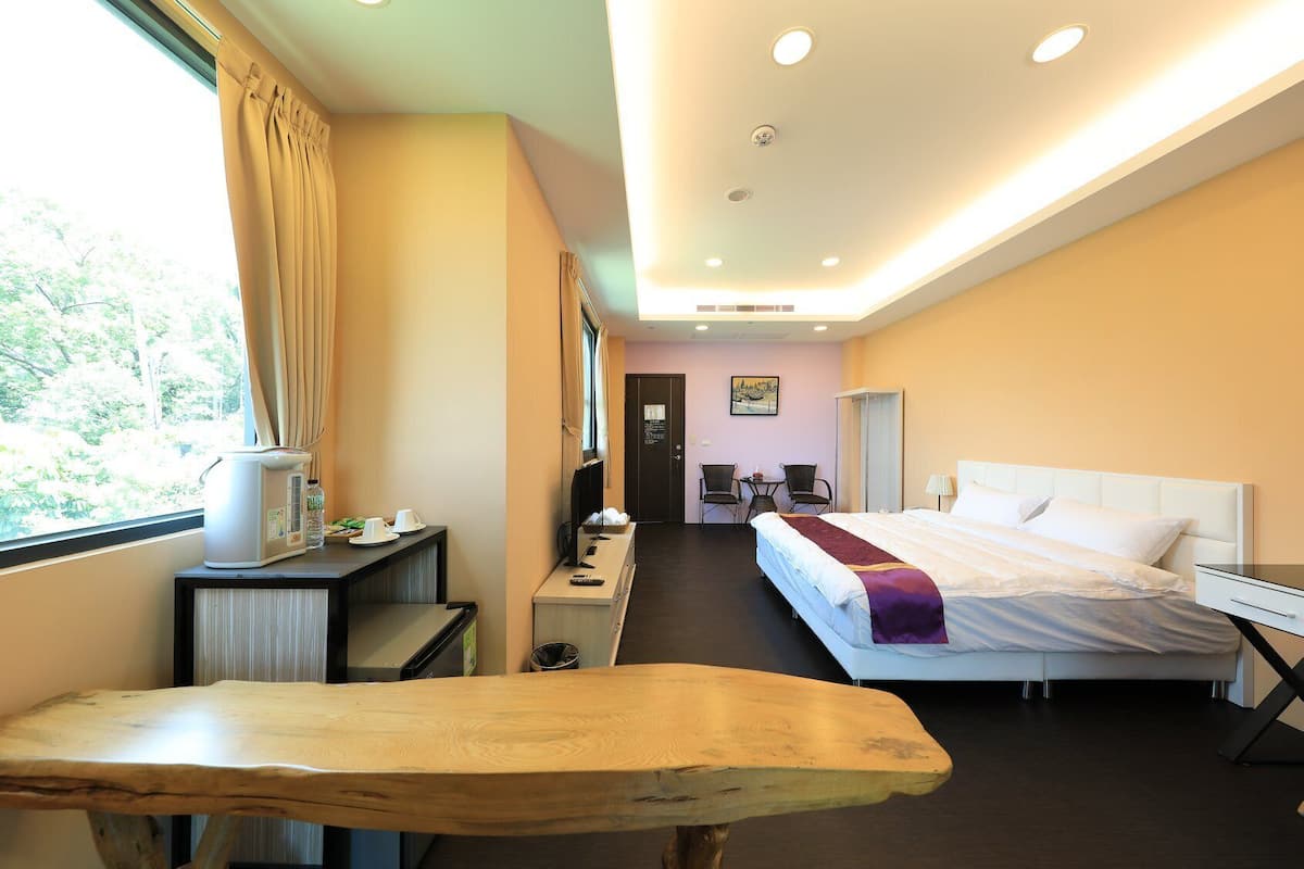A hotel room with a bed, bedside table, desk with a computer, and a window with curtains.