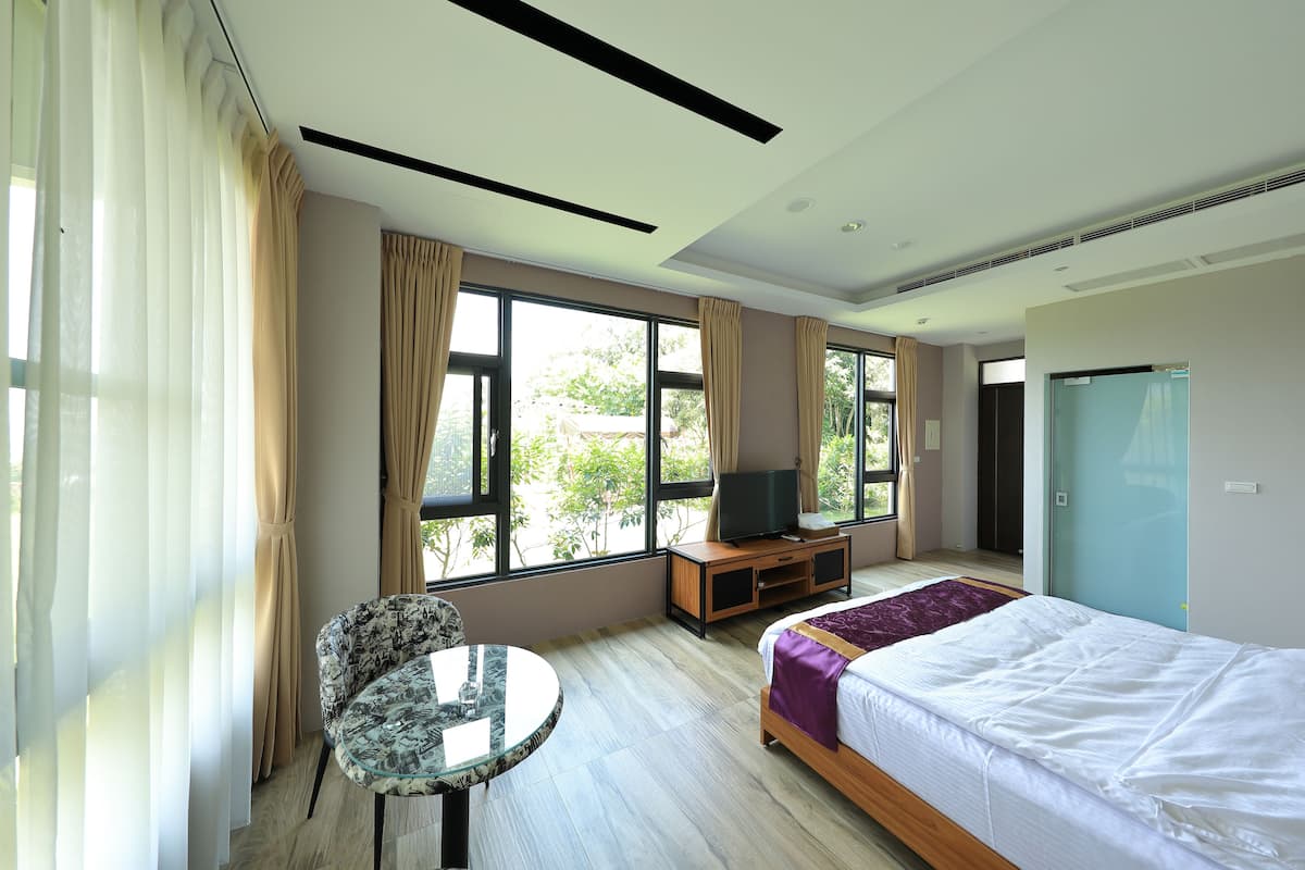 A hotel room with a bed, a TV on a wooden stand, a round table, and large windows with curtains.