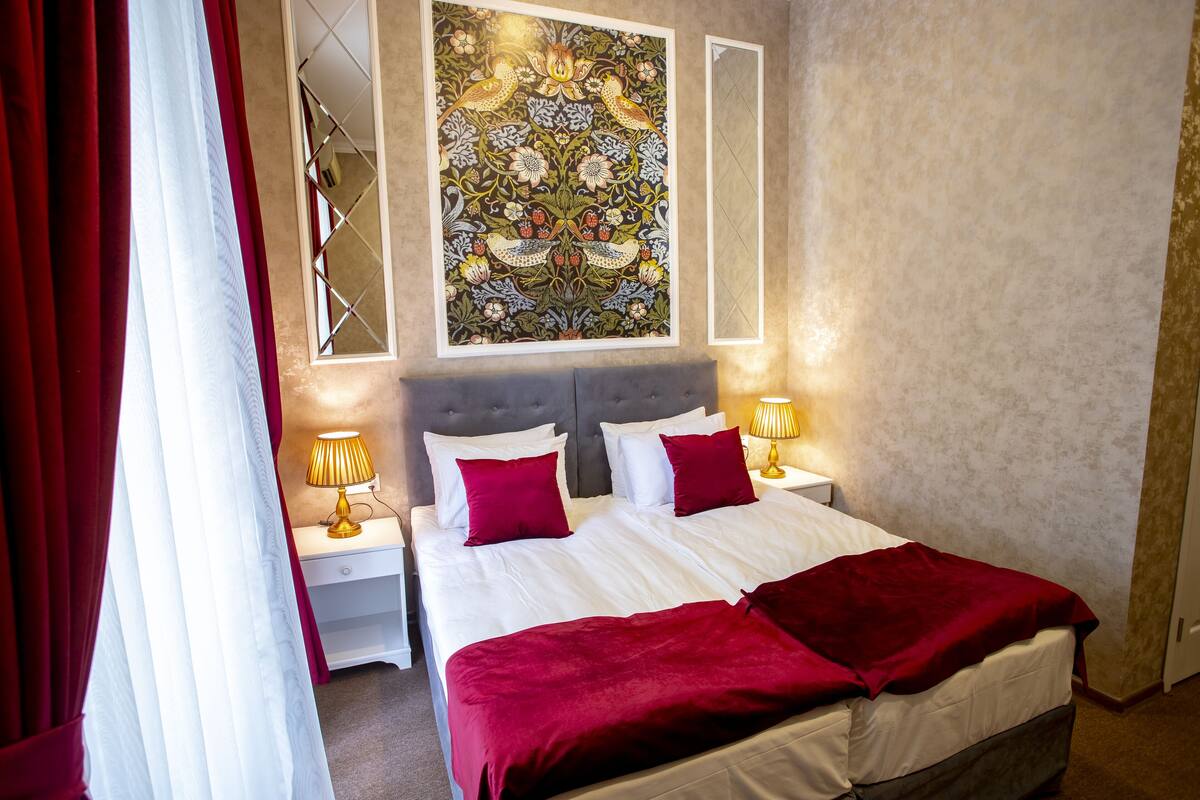 Un lit bien fait, avec une literie blanche et rouge, deux tables de chevet avec des lampes, une grande œuvre d’art florale accrochée au mur et un rideau rouge partiellement tiré.