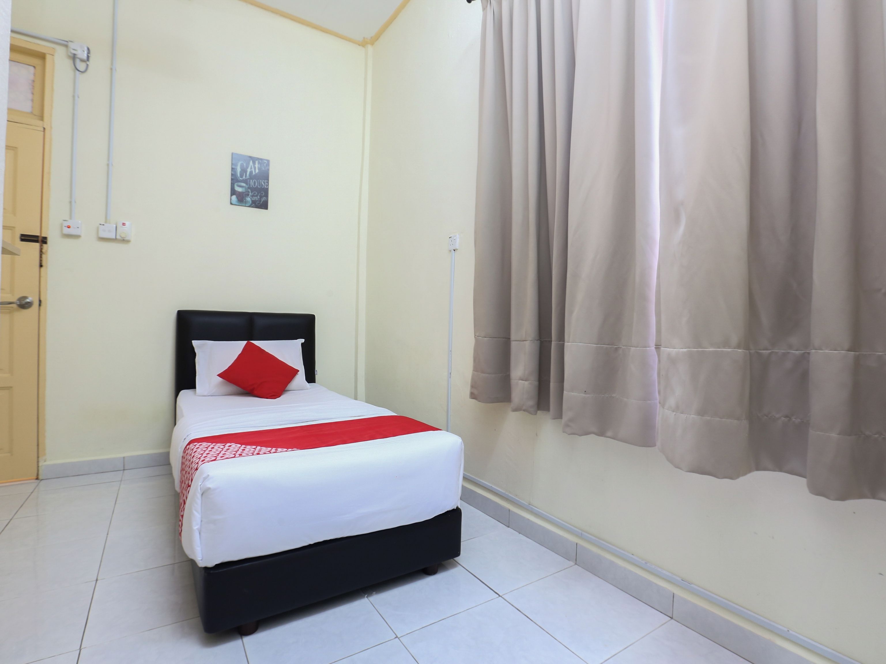 A hotel room with a bed, a red pillow, a white bedspread, a beige curtain, and a small sign on the wall.