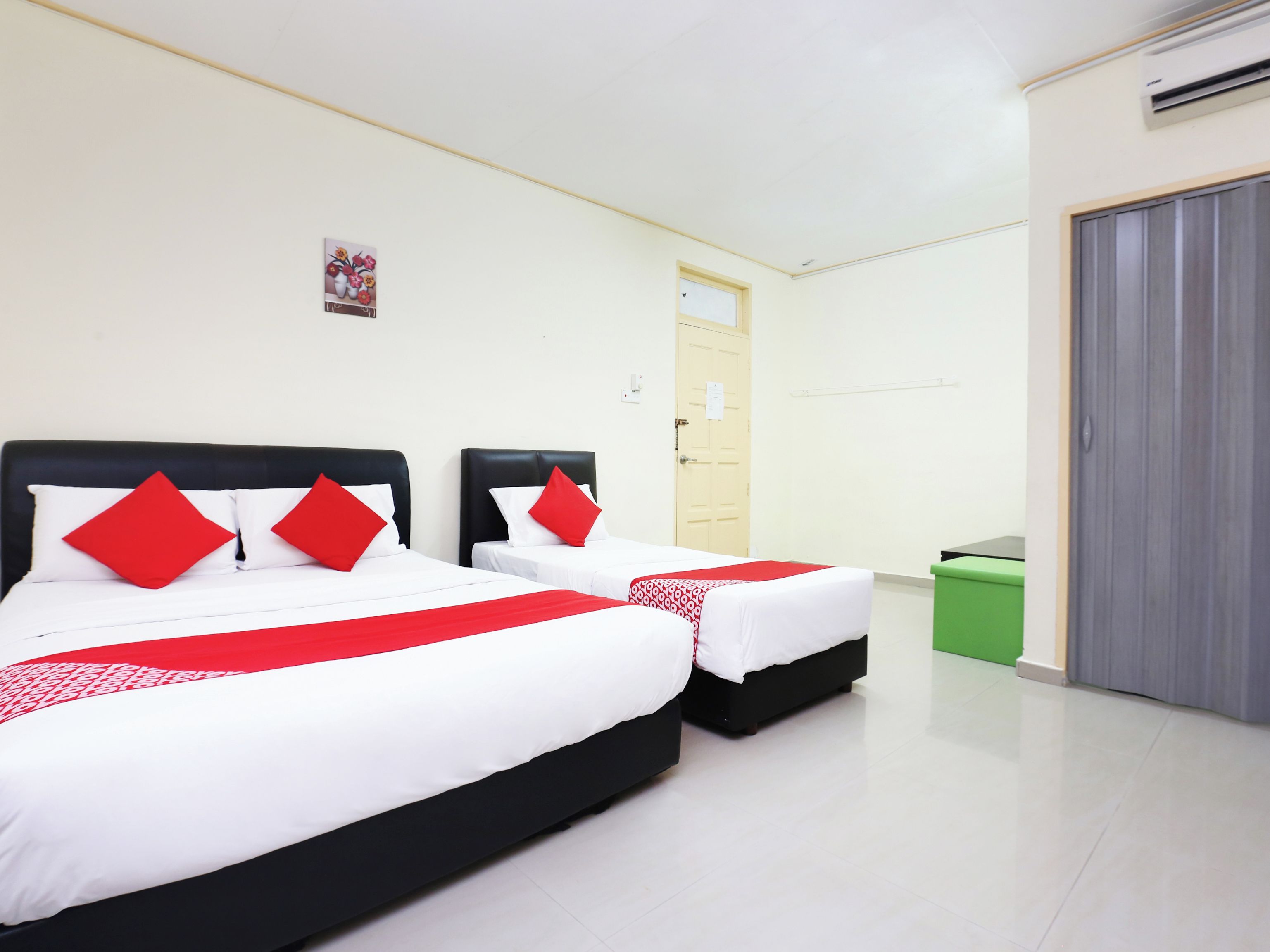 Habitación de hotel con dos camas, cada una con cabecero negro y ropa de cama blanca con detalles rojos. Hay un cubo de basura verde y una puerta cerrada en la habitación.