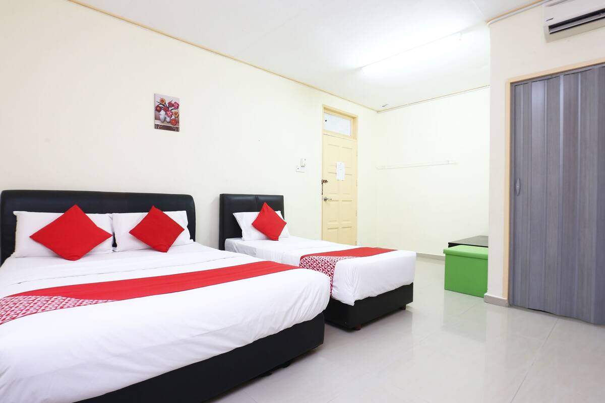 Habitación de hotel con dos camas, cada una con ropa blanca y almohadas rojas, un cubo de basura verde y una puerta gris.